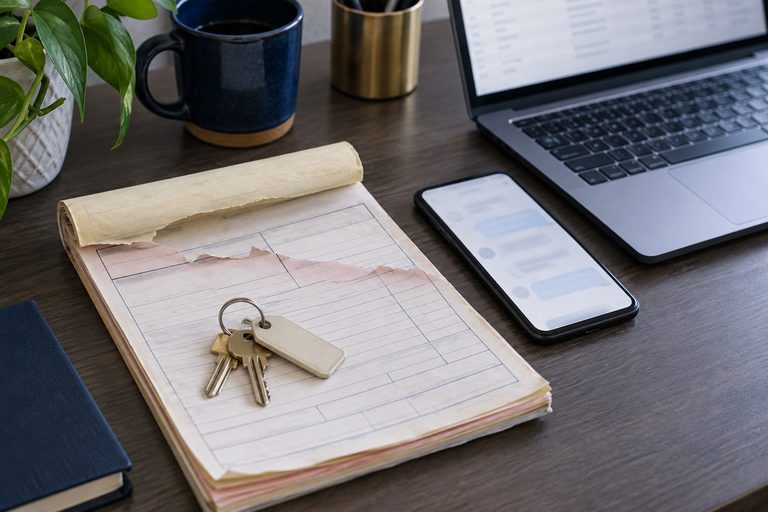 One repair living in three places at once: a carbon-copy work-order pad with a ring of tagged keys on it, a phone showing an indistinct message thread, and a laptop showing an indistinct table.